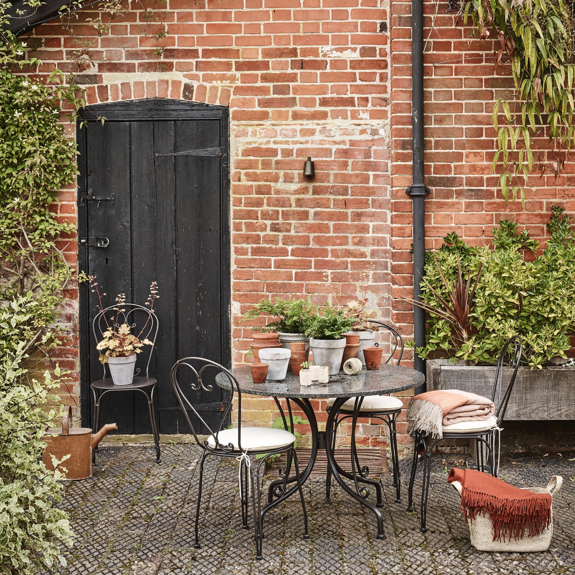garden with paved area near outbuilding door, bistro set with blankets and cushions, pots on table, planter to the right, basket with blankets