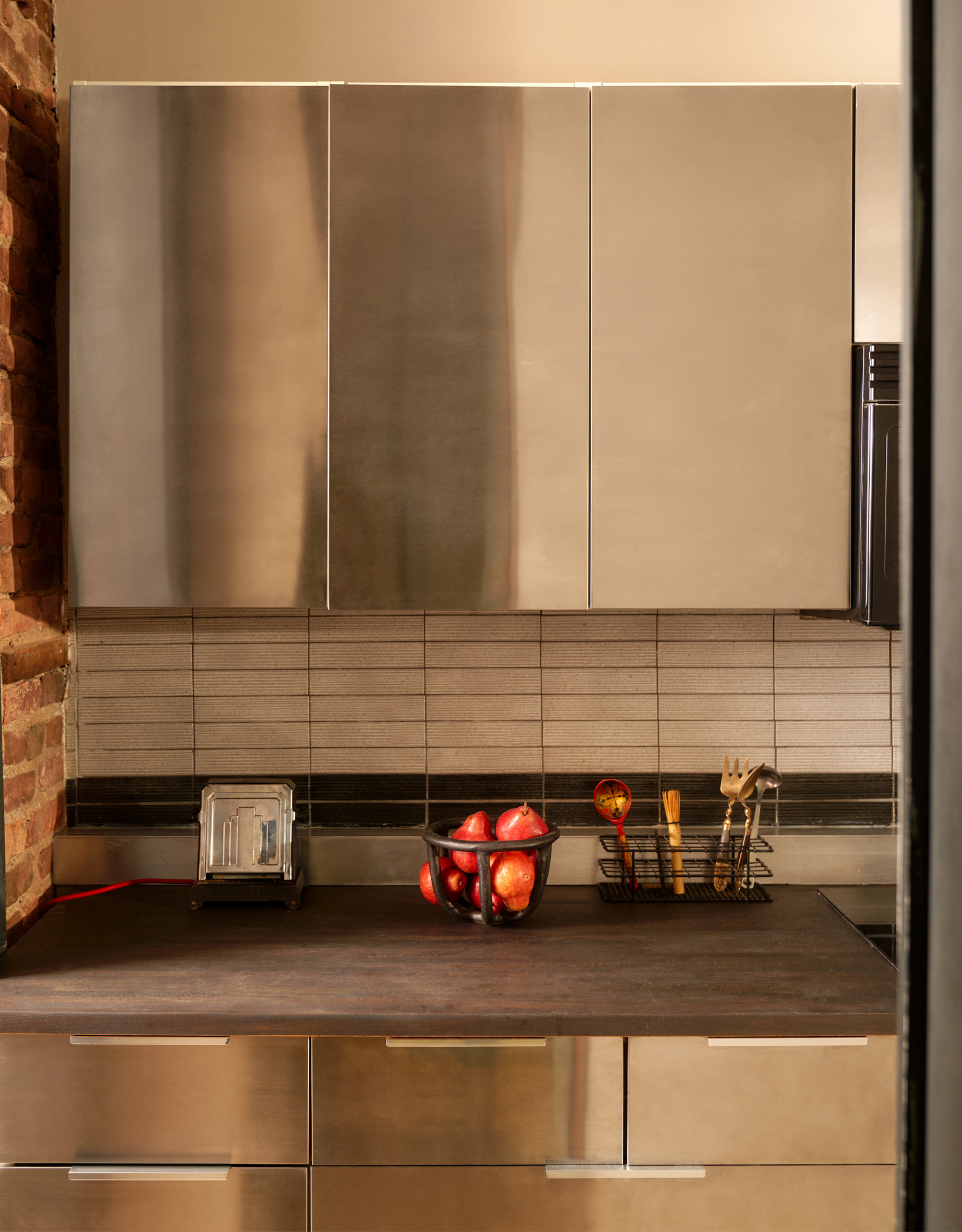 A NYC loft kitchen with stainless steel cabinets, a tiled backsplash, and a wood top with a bowl of fruits