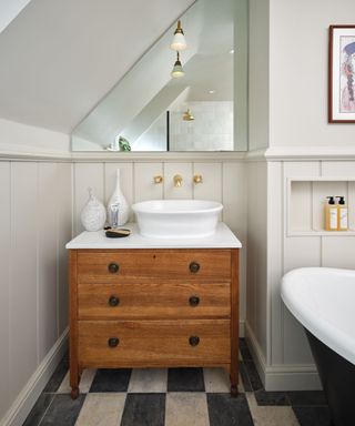 A bathroom with a vintage aesthetic featuring a wooden chest of drawers repurposed as a vanity with a white countertop and an oval vessel sink. Gold taps are mounted on the wall above the sink. A large mirror with a sloped upper edge reflects the space