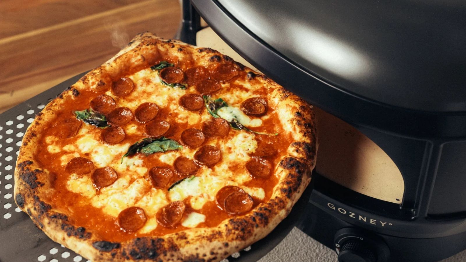 A cooked pepperoni pizza coming out of a black slimline pizza oven on a perforated pizza slice. 