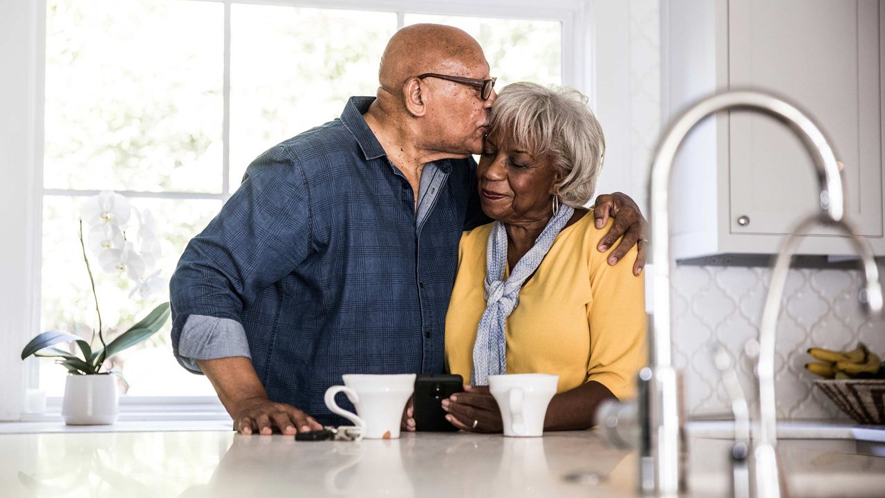 An older man kisses his wife&amp;#039;s forehead.