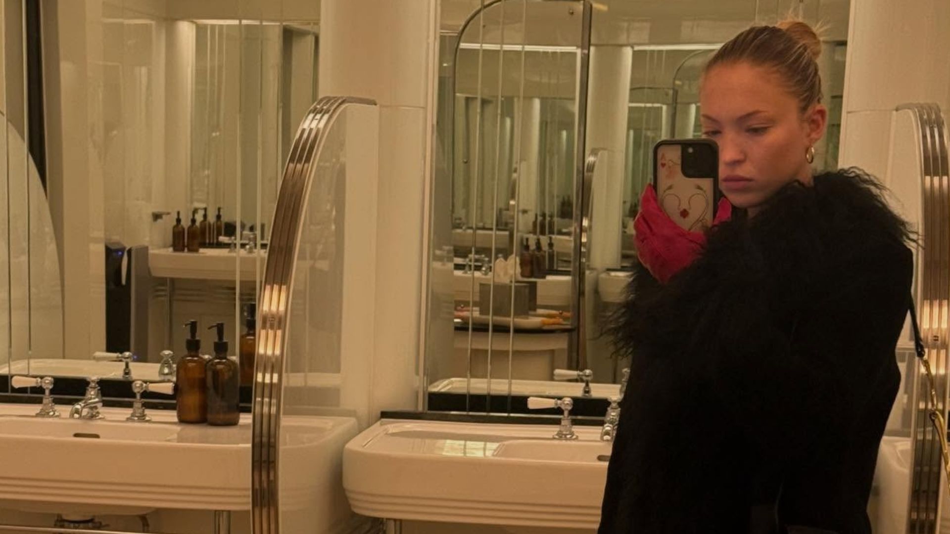 Lila Moss takes a mirror selfie in a bathroom wearing bootcut jeans with rounded-toe heels boots and a cur-trimmed jacket.