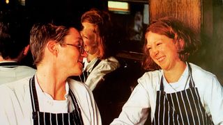 Fergus and Margot staring at each other. Both wearing chef whites and a black and white apron.