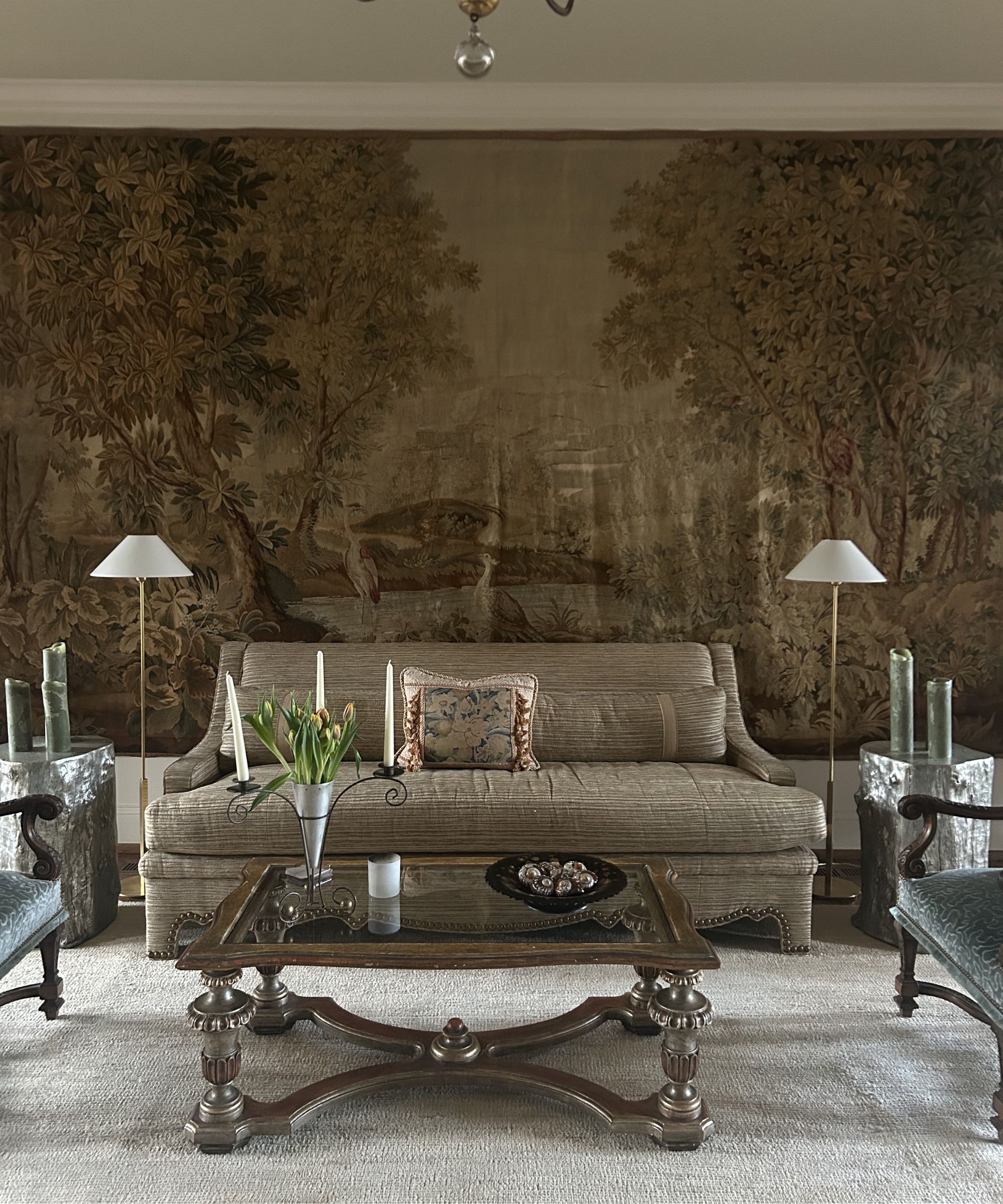 Living room with antique tapestry wall hanging, a beige carpet, a brown loveseat with a tapestry pillow, an ornate glass coffee table and two floor lamps on either side of the sofa.
