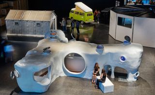 View from above of the 'Design at Large' exhibition featuring a white cave-like structure, a green 1960s Volkswagen Campervan, a multisided light grey structure with a window and flat roof and a white structure with a pitched roof. There are also a number of people around the exhibition pieces