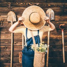 A woman in a straw hat and overalls playfully hides her face behind the hat while standing against a rustic wooden wall. The themes of relaxation, and the simple joys of country life.