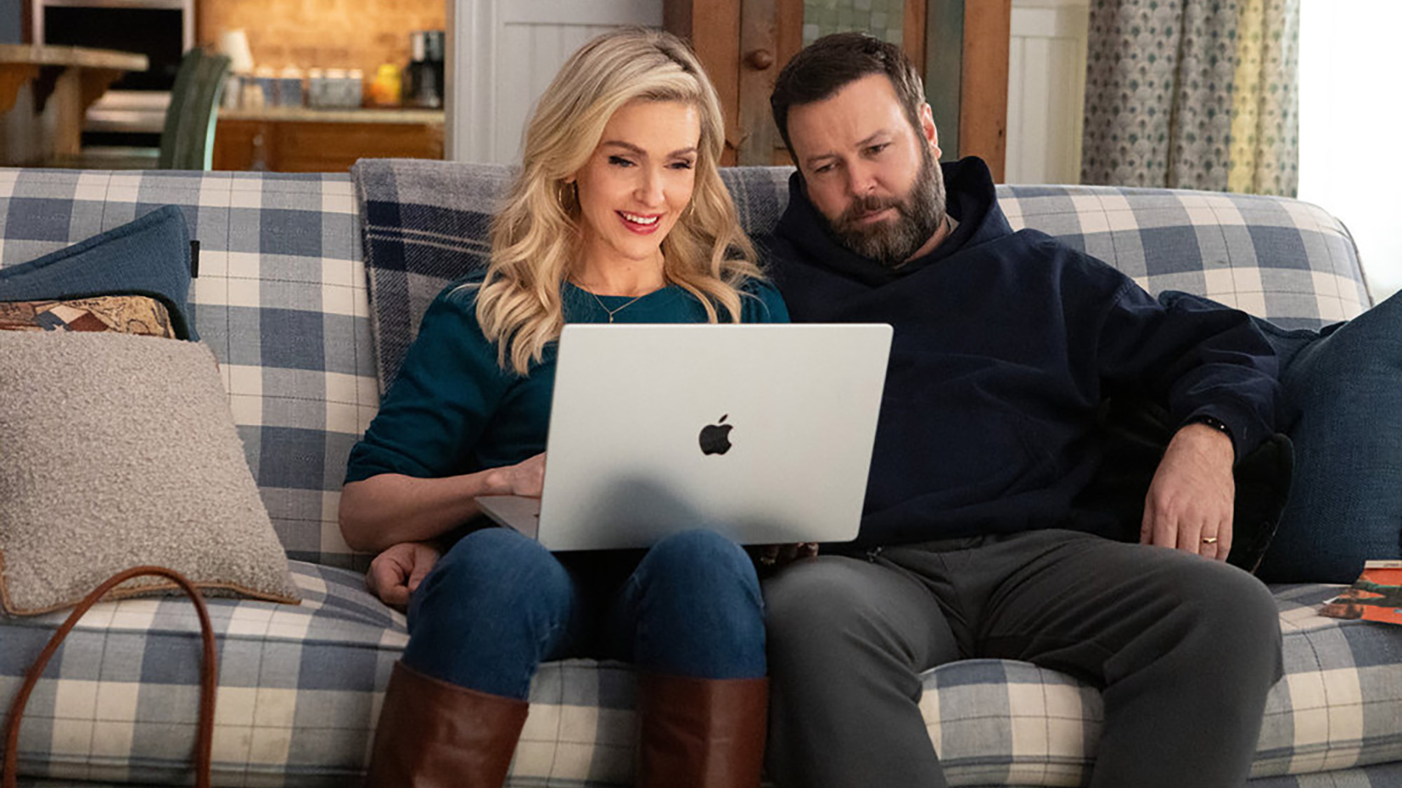 Jenn Lyon and Taran Killam sit on a coach and watch cheerleading footage on a laptop in Stumble.