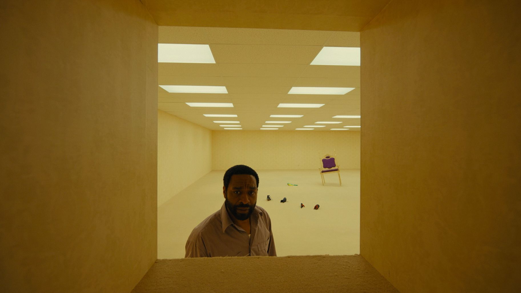 A pale yellow film set with textural wallpaper, minimal decor, and people wandering through it.