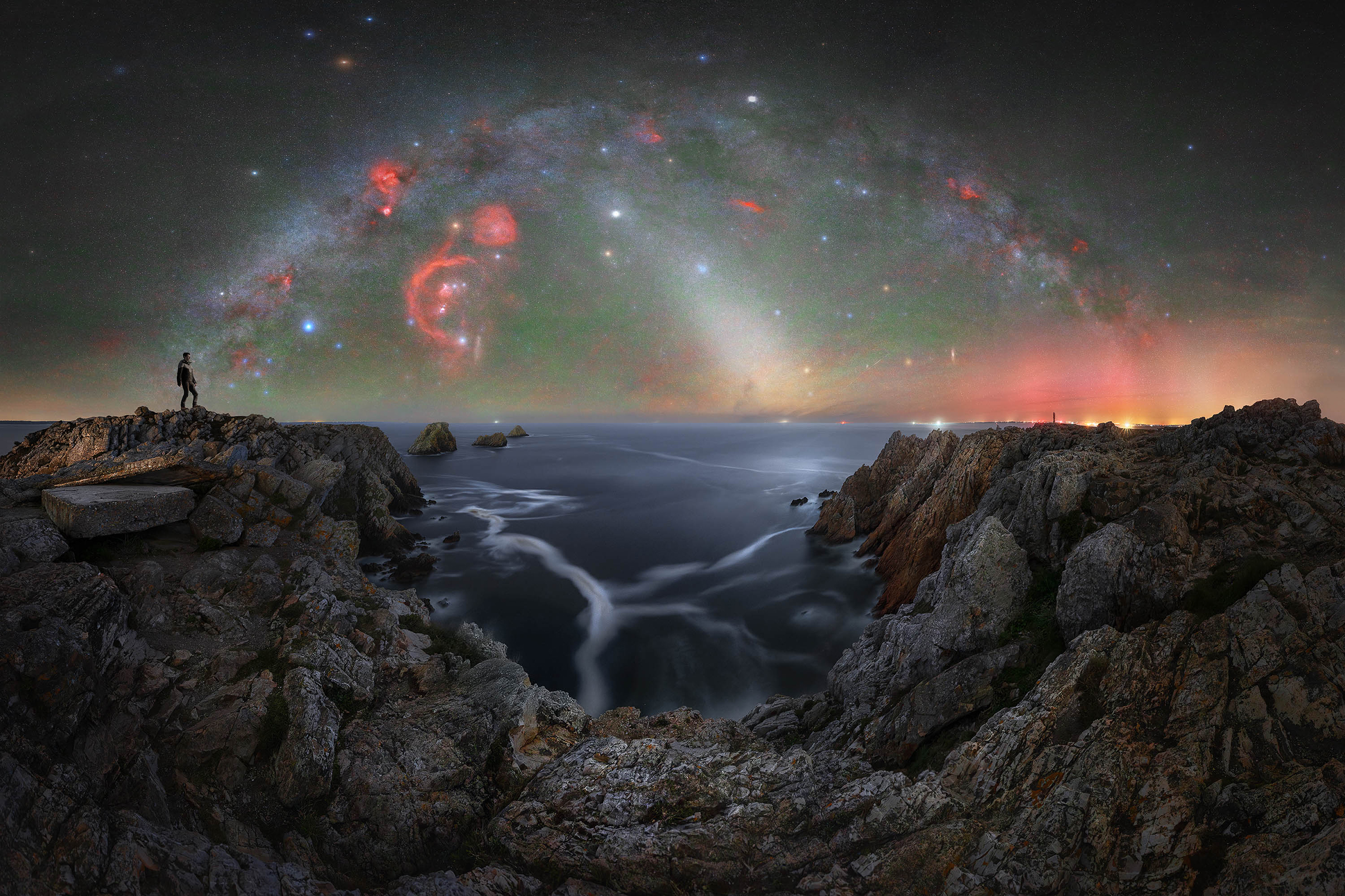 Person stands on rocky cliffs overlooking a calm sea under a vibrant, star-filled night sky with colorful nebulae and galaxy arches