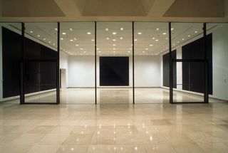 The room has beige tiles with white walls. Through an all-glass divider, we see square black canvases on the walls.