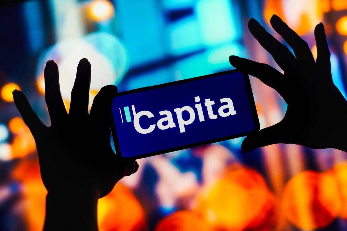 Capita logo appearing on a smartphone that&#039;s being held out in the fingertips of two hands, the arms of which are visible, appearing form each side of the frame, all silhouetted