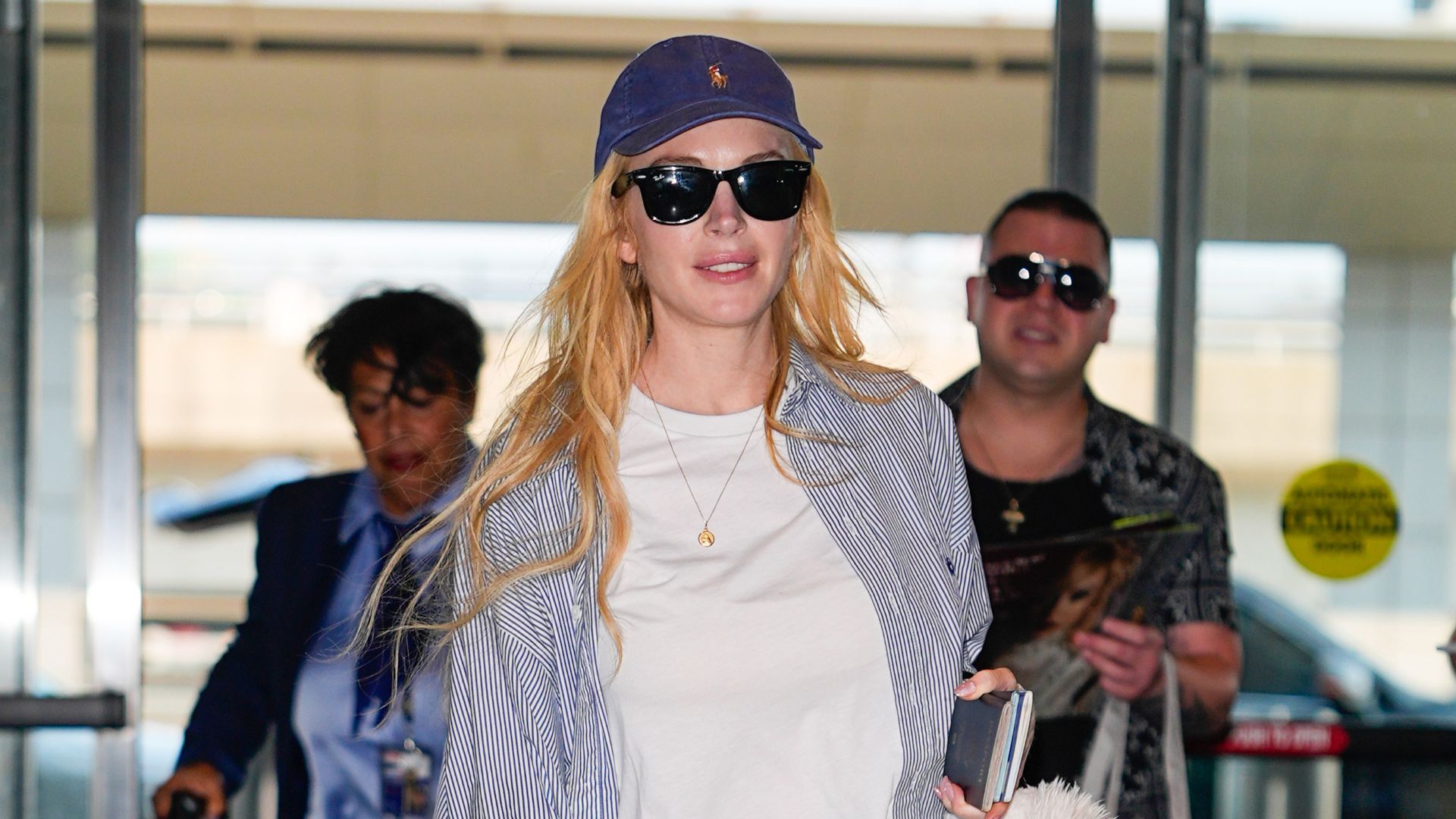 Lindsay Lohan walks through the airport wearing dark navy leggings with a white t-shirt and a striped shirt and white trainers.