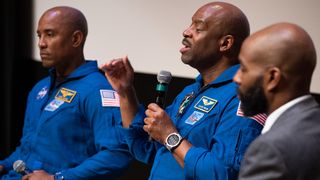three speakers in a row on a stage. the two on the left are astronauts and wear astronaut suits. one holds a microphone. the last person on the right wears a suit