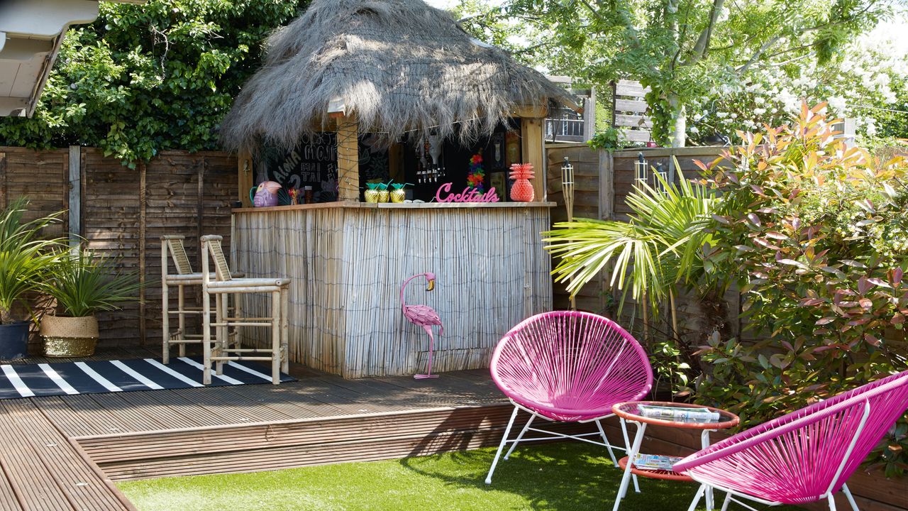 Garden with a tiki bar with palm roof, bar stools and decking with astroturf lawn and pink chairs