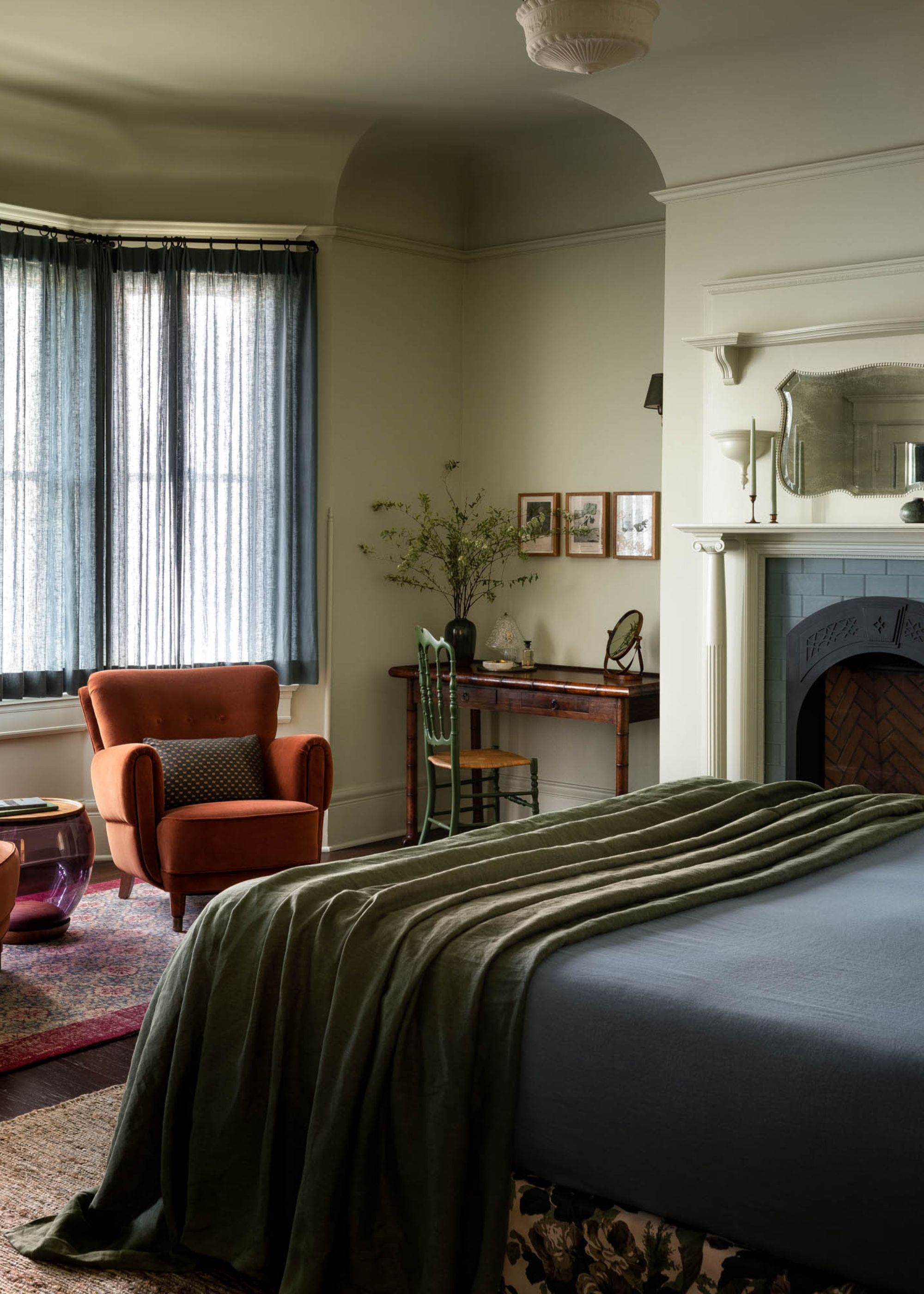 A bedroom with light green color-drenched walls, dark navy bedding, a bay window with sheer window treatments, and a red armchair with a burgundy rug beneath.