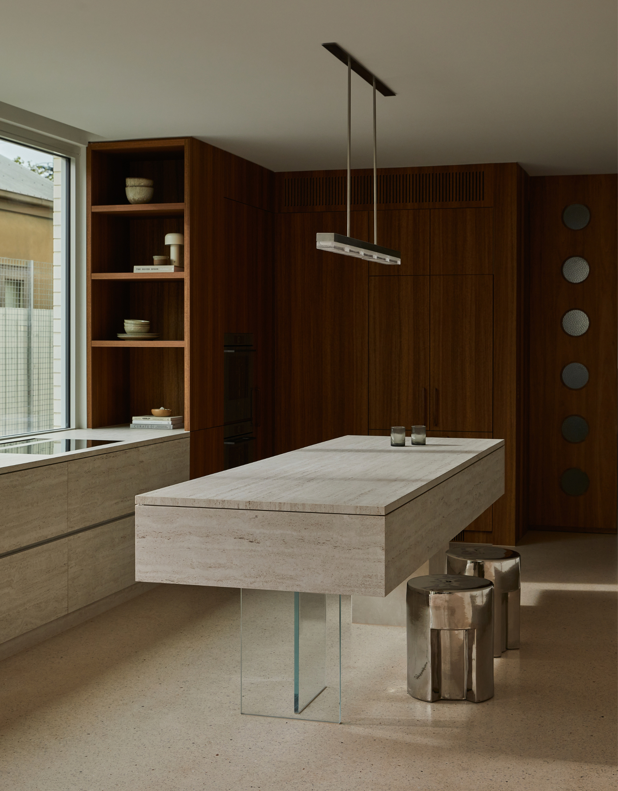A minimalist kitchen with a stone and glass island, a pair of chrome stools, and an overhead light