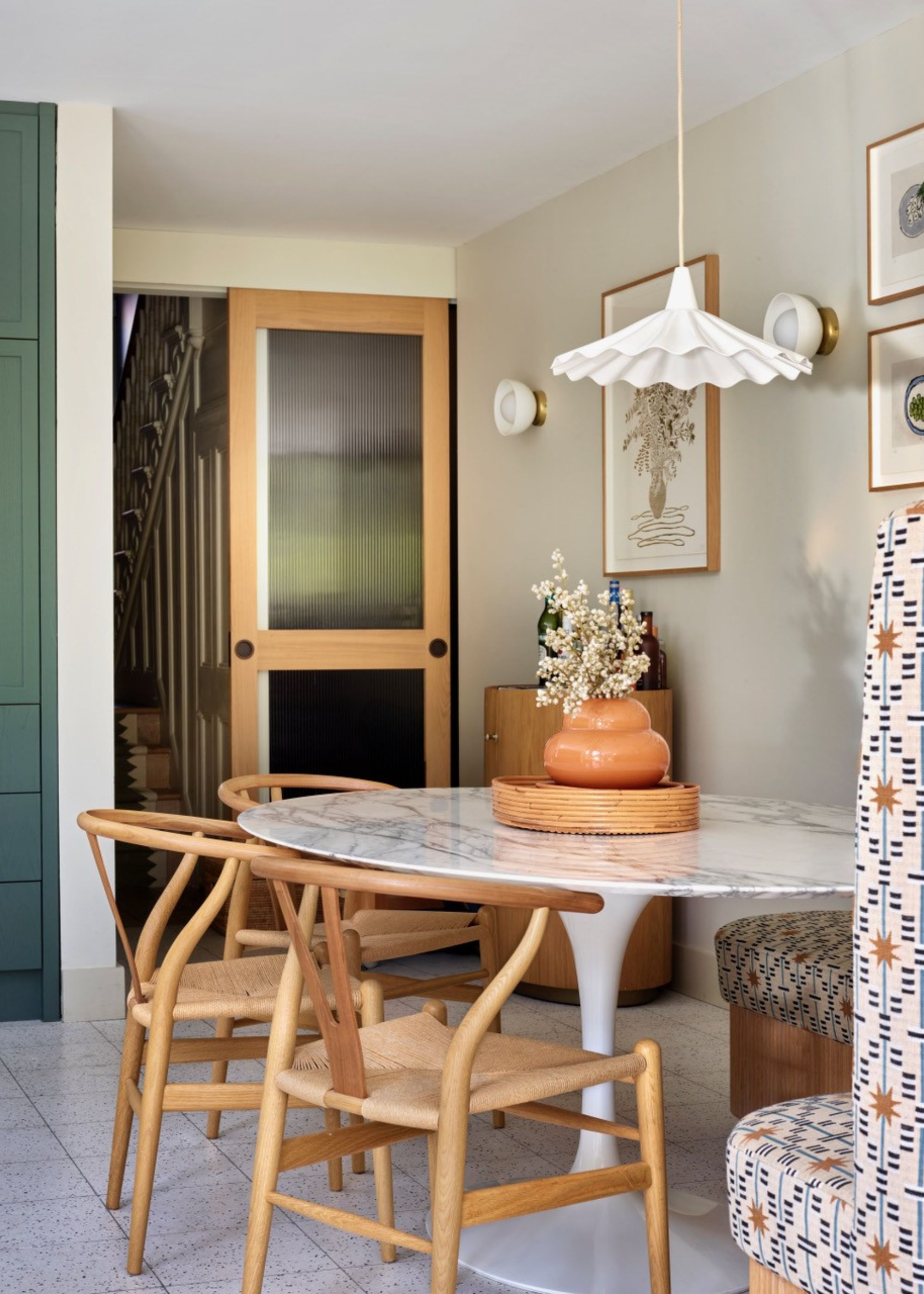A modern kitchen extension with a patterned, curved banquette seating area just out of view by a round marble dining table with wooden curved back chairs and a wave pendant light above