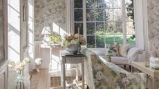 Floral living room with floral wallpaper, furniture, light wood flooring, and large windows with a green garden seen beyond.