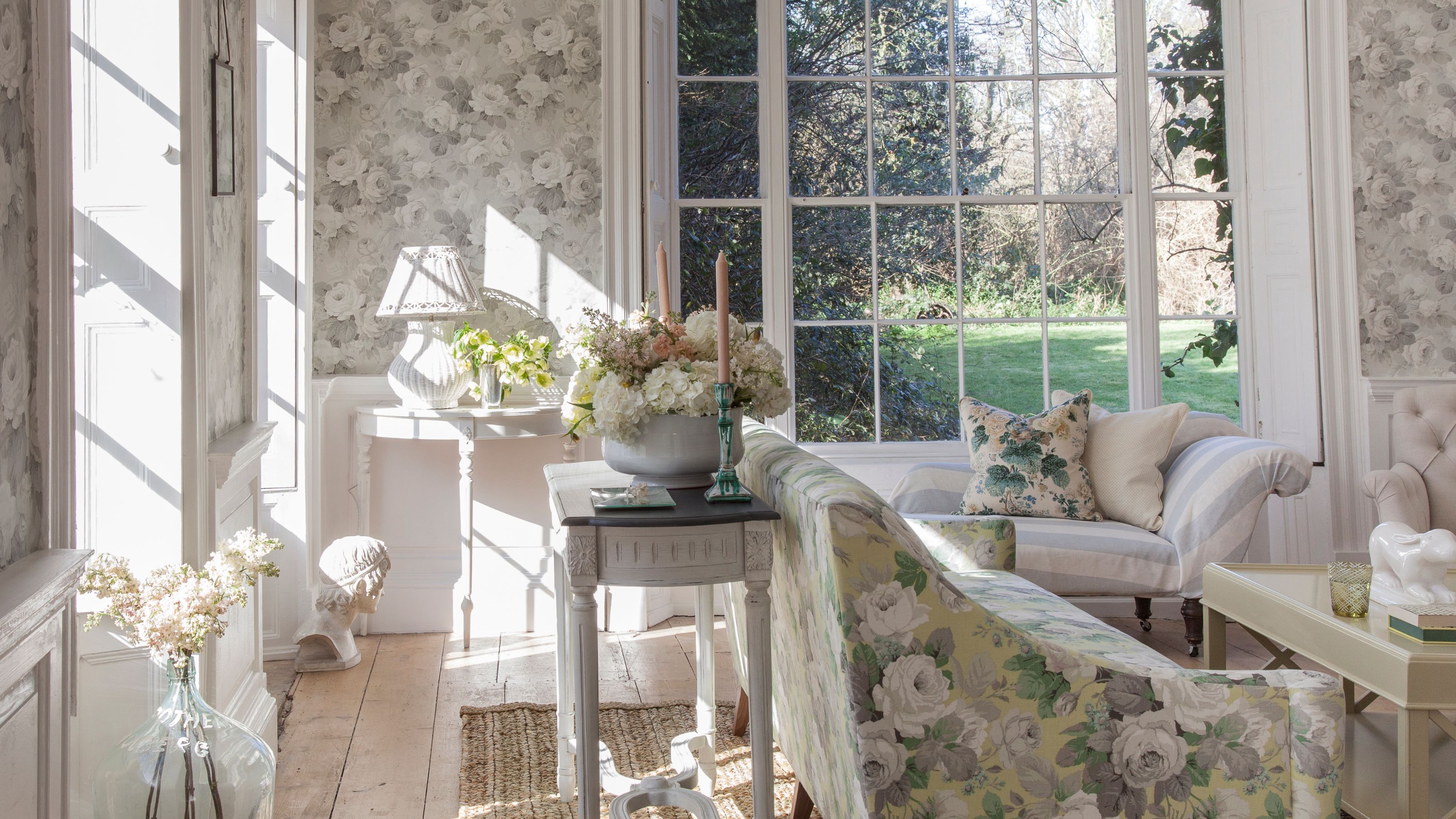 Floral living room with floral wallpaper, furniture, light wood flooring, and large windows with a green garden seen beyond.