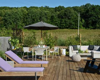 A decking area with purple loungers, L-shaped sofa and small table and chairs opening onto a field and trees.