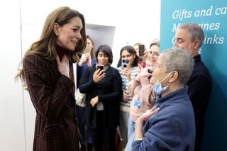 Kate Middleton wearing a burgundy plaid coat putting her hand on her chest while talking to a woman wearing a surgical mask pulled under her chin at a hospital with a group of people recording on their phones behind her