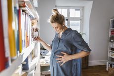 Woman looking at her pregnant belly next to a bookshelf at home