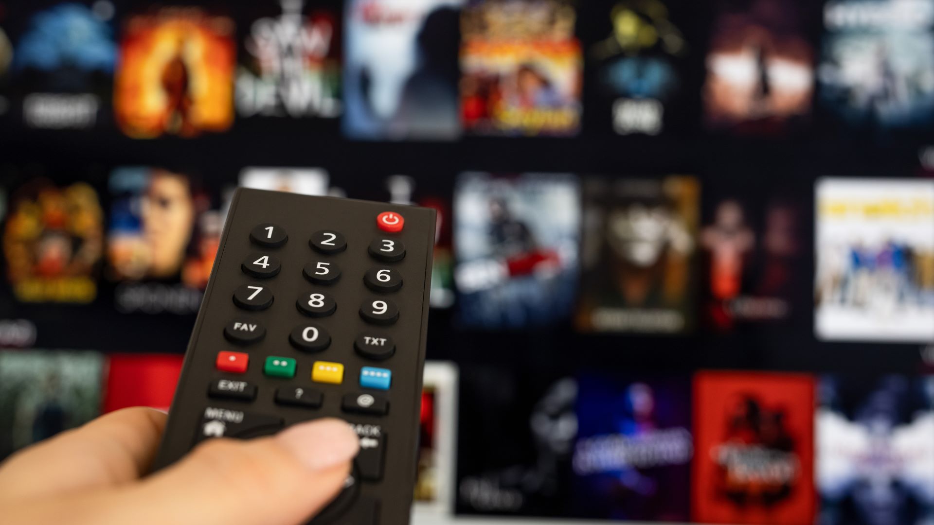 A person holds a TV remote in the foreground. In the background is Netflix's interface, with many movies. The background is heavily blurred