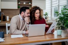 Couple at home managing personal finances, reviewing financial documents and monthly budget on a laptop, calculating bills