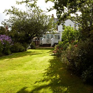 Large garden laid mostly to lawn with trees and shrubs to its side, and a house in the distance