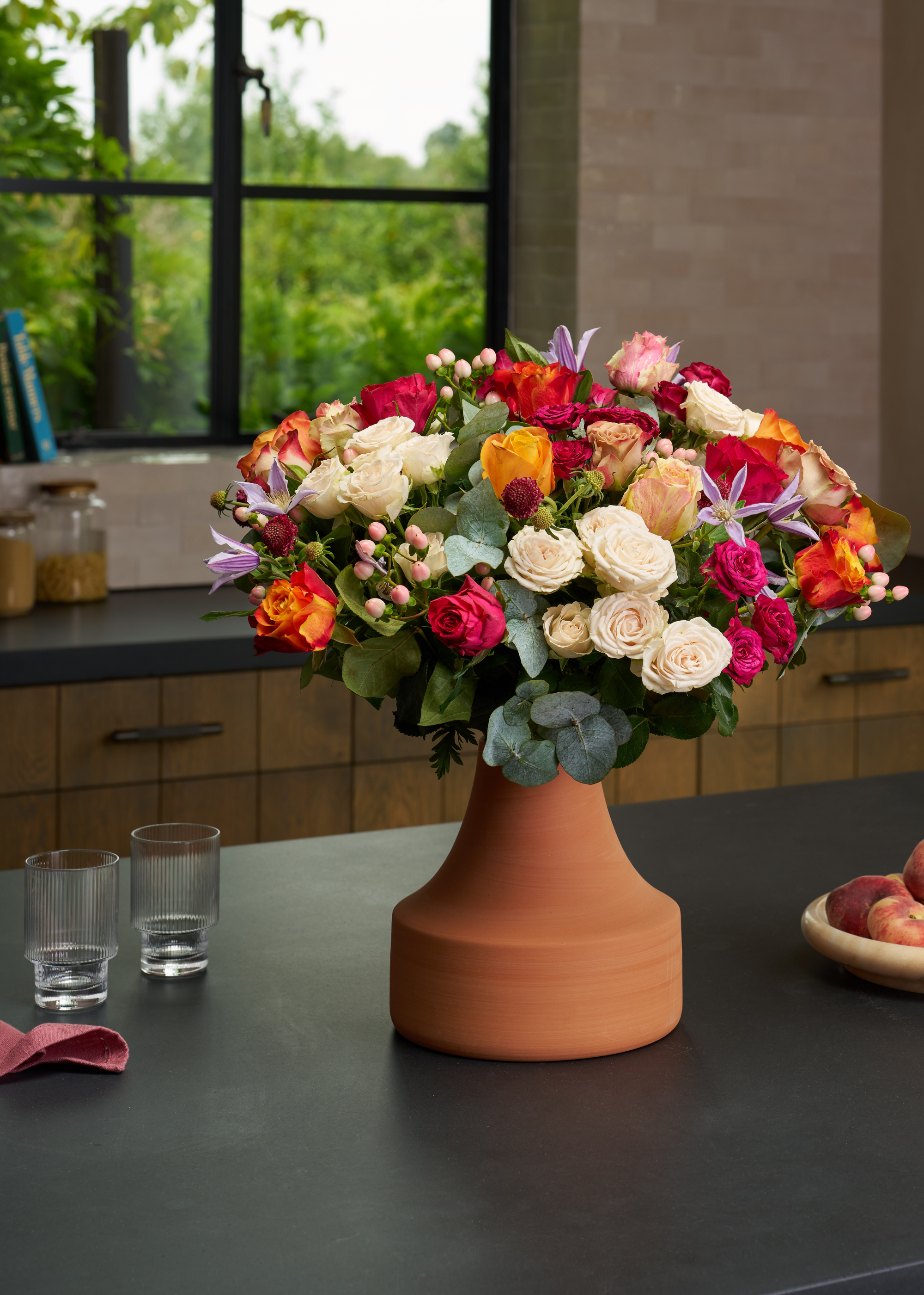 A terracotta vase of colorful flowers including roses and eucalyptus on a black kitchen island beside a bowl of peaches, a pink cloth and a pair of ribbed drinking glasses