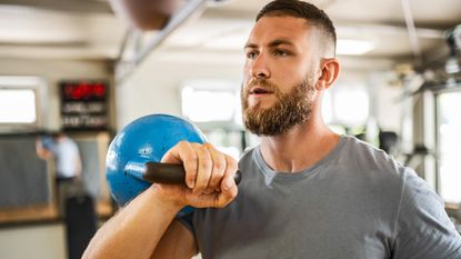 A man exercises in a gym with a kettlebell. He is wearing a t-shirt and has a kettlebell hooked over his right hand, which rests on his shoulder. Behind him we see gym apparatus.