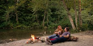 Mel and Jack sitting next to a river as a small logfire burns in one of the Virgin River locations