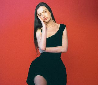 Cazzie David poses in front of a red backdrop wearing a Mugler asymmetric black dress.