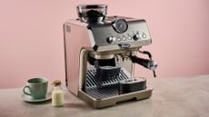 The Delonghi Specialista Arte Evo sits on a stone-effect surface in front of a pink backdrop. It has a green cup and saucer, and a small cork-topped bottle of milk to its left.