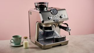 The Delonghi Specialista Arte Evo sits on a stone-effect surface in front of a pink backdrop. It has a green cup and saucer, and a small cork-topped bottle of milk to its left.