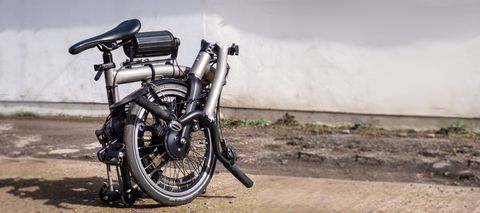A black and silver folded Brompton T-Line, upgraded with the Cytronex e-bike conversion kit, sits on a rocky surface