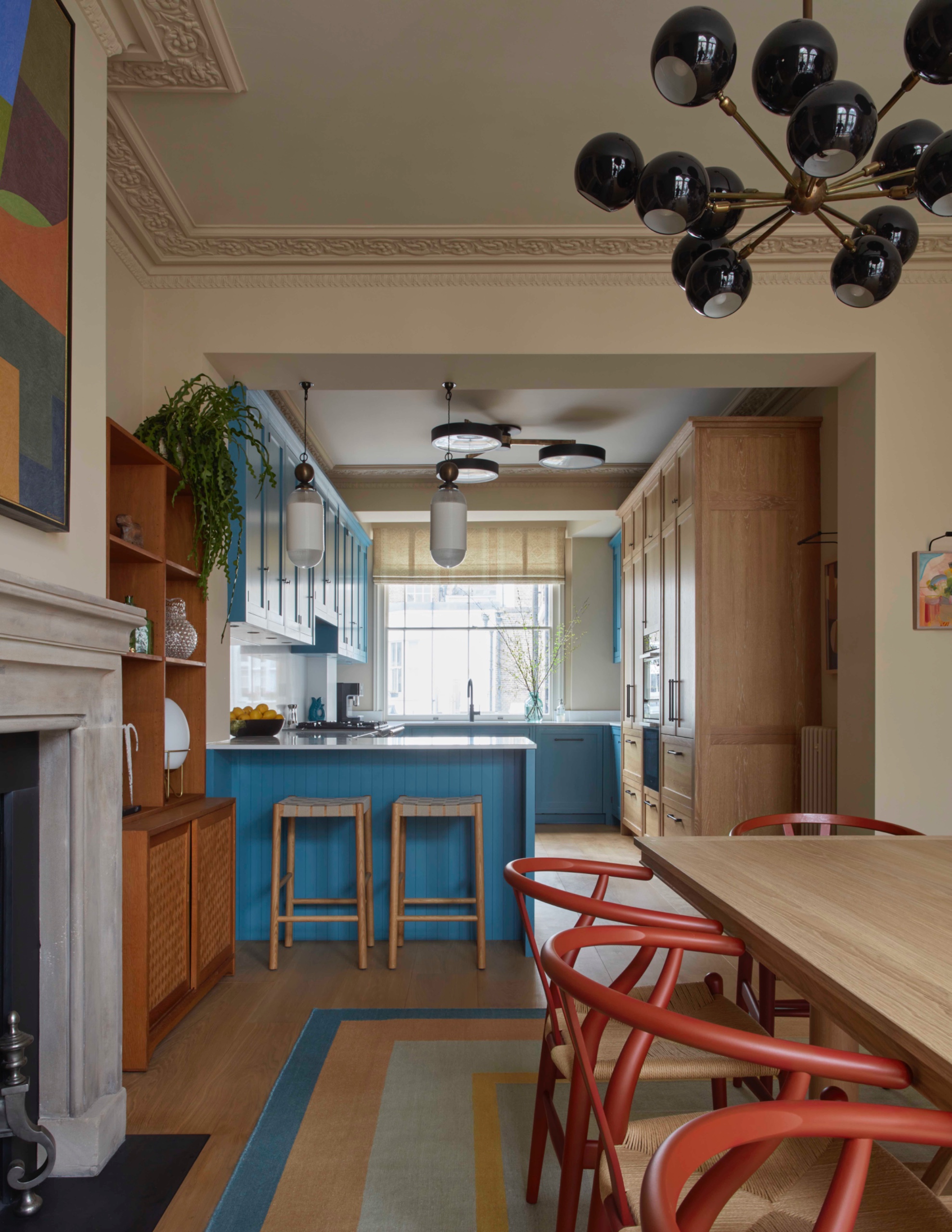 Dining room with a wooden table, red chairs and a fireplace, looking through a wide opening into a kitchen with blue cabinetry, tall wood cabinets and a peninsula with two stools.