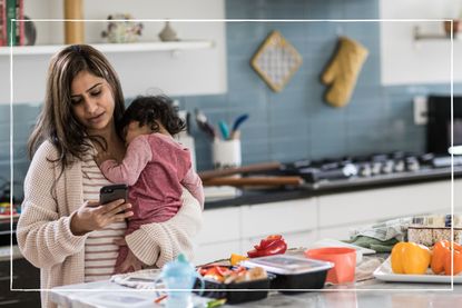 Mother holding daughter in the kitchen while also looking at her phone