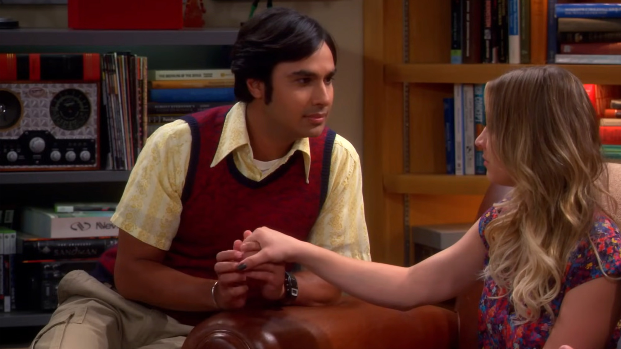 Raj in a button down and sweater vest holding Penny's hand and staring straight into her eyes on The Big Bang Theory couch. 