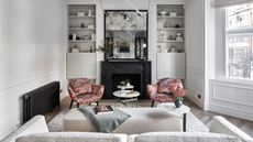 A white living room with white rug, white couch, white footrest, pink and red patterned chairs, a black radiator, and white shelving around a black marble fireplace with a black mirror above.