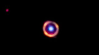 A blue dot surrounded by an orange rings on a black background. In the left corner, a red dot glows.