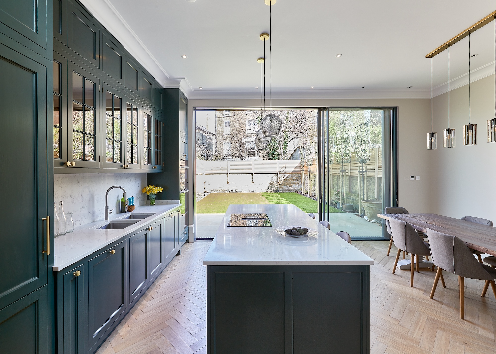 A dark green kitchen with an island in the middle and pendant lights hanging above the island. There are also glass fronted cabinets with kitchen accessories inside as well as a large dining table on the right side of the kitchen