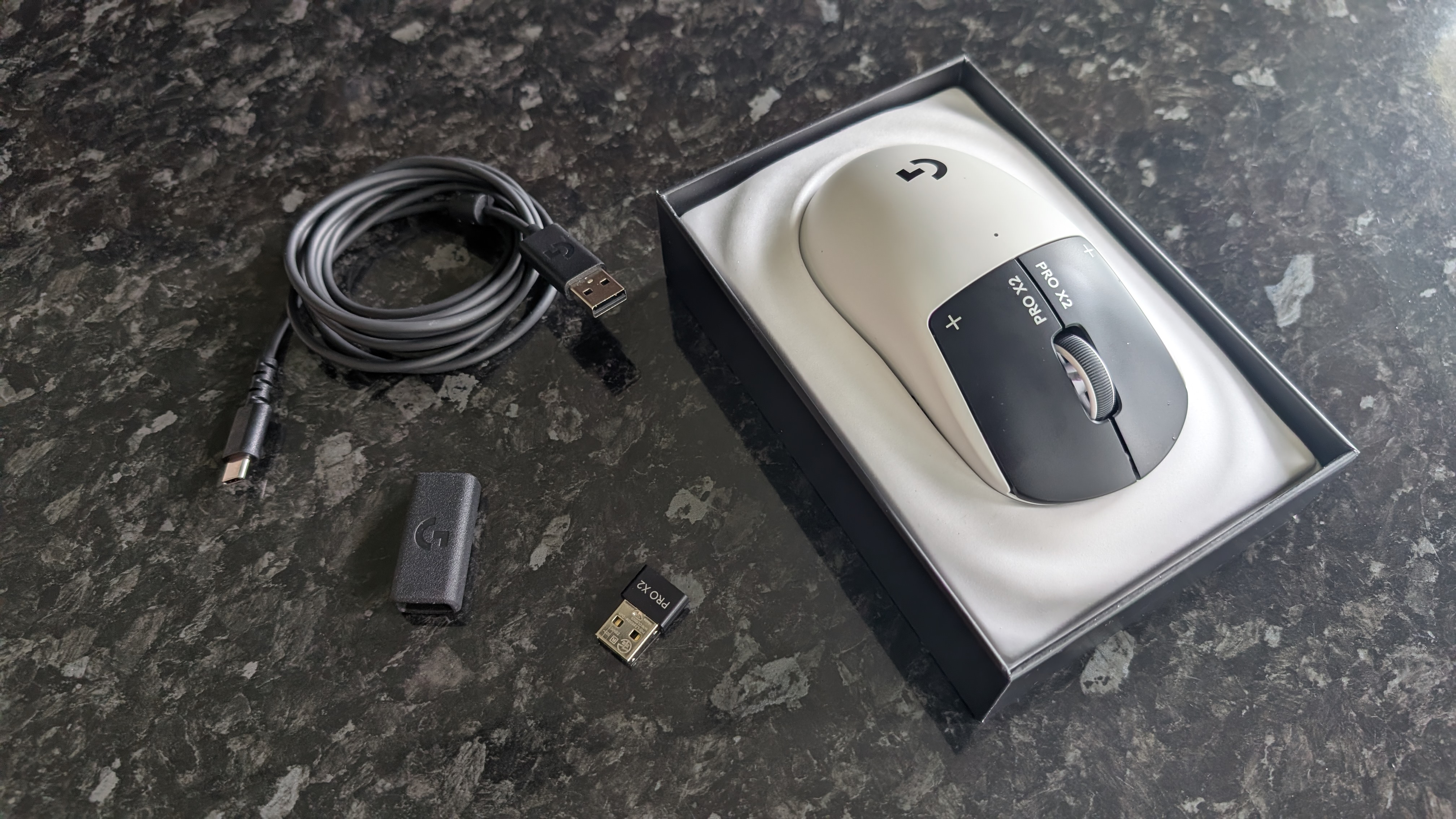 The Logitech G Pro X2 Superstrike pictured on a black marble surface, in the box, with wireless dongle, adapter, and USB cable.