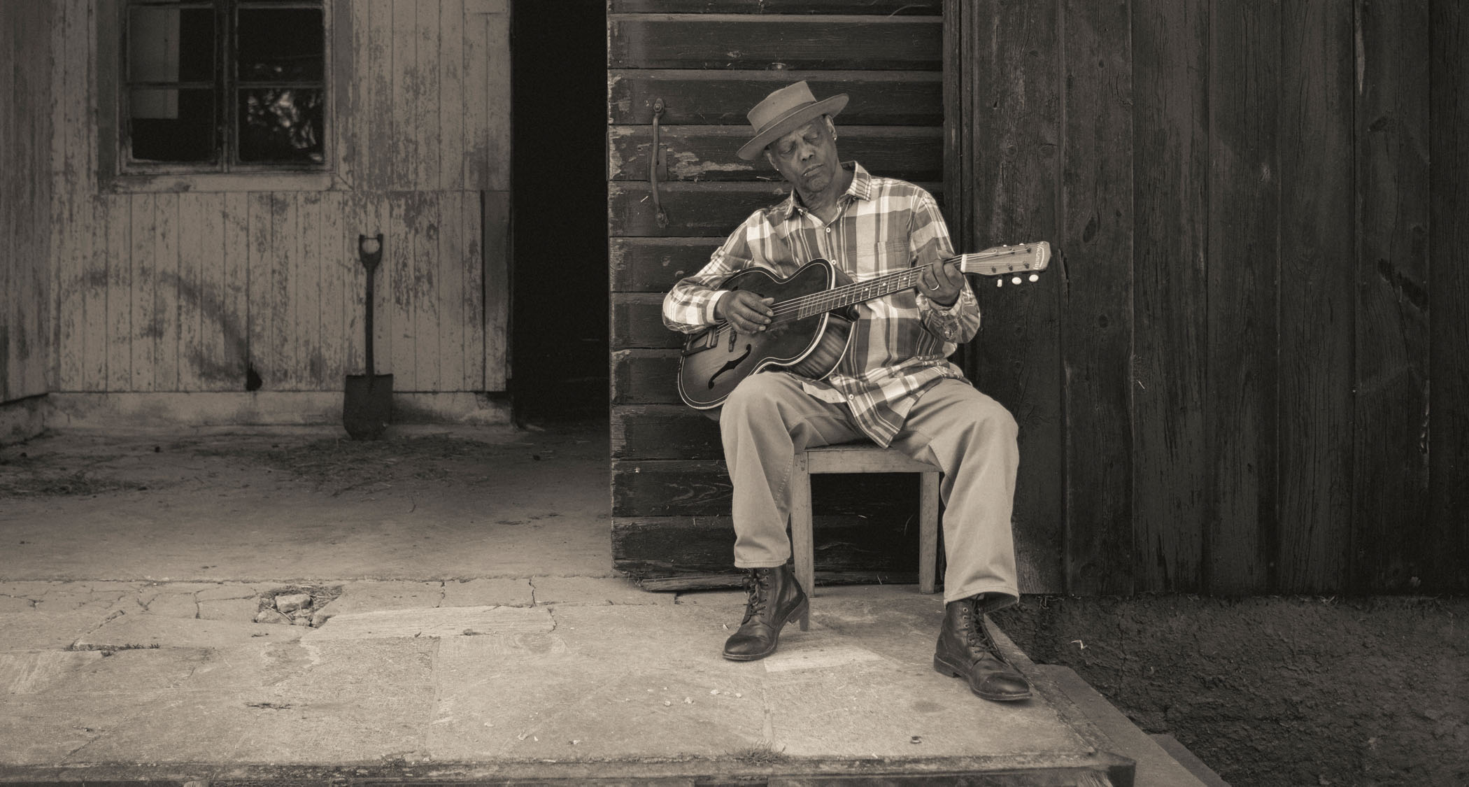 A black-and-white press shot of Eric Bibb playing some acoustic blues on a chair outdoors