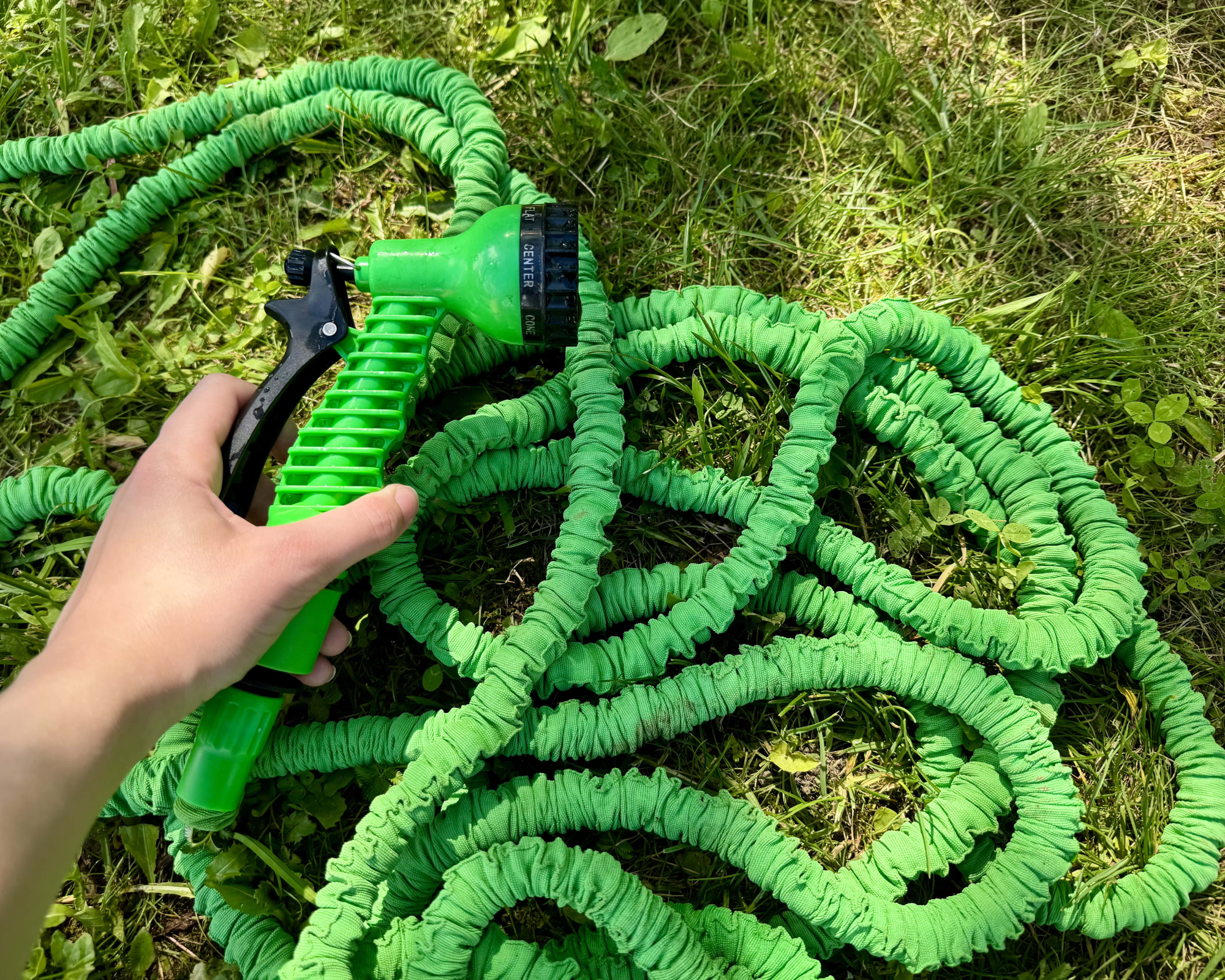 Hand holding green expandable garden hose with spray nozzle on grass in summer garden