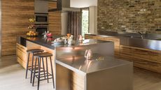 A modern kitchen with exposed brick walls and wooden cabinets. In the center is a stainless steel island counter with black bar stools, and to the right is a stainless steel countertop with a sink.