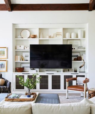 Inviting white living room with dark wood accents, large storage and display unit with tv, leather chair, blue textured coffee table and cream sofa