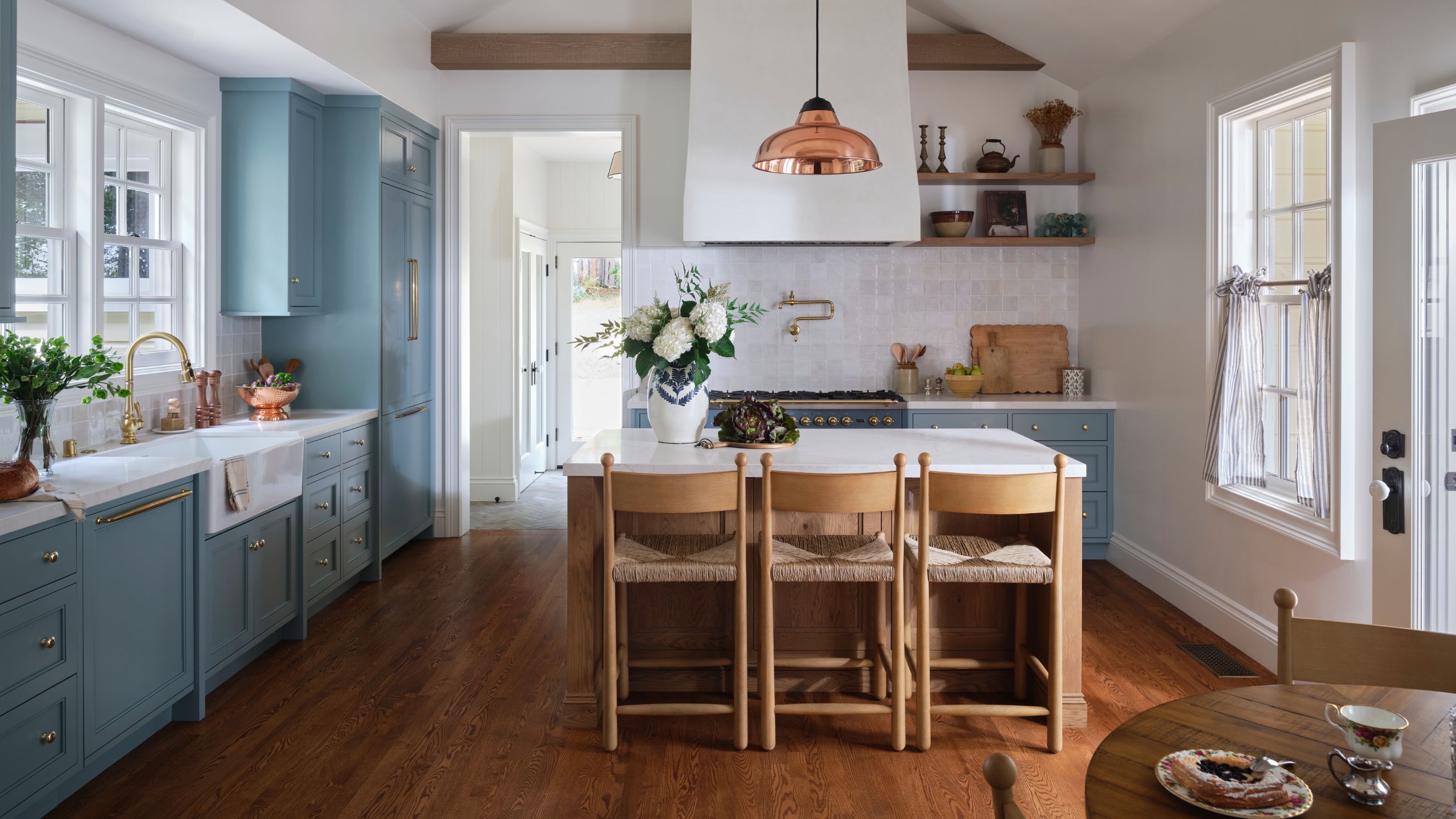 a white and bright kitchen with ceiling beams and light blue cabinetry, a small island with oak stools and a copper pendant