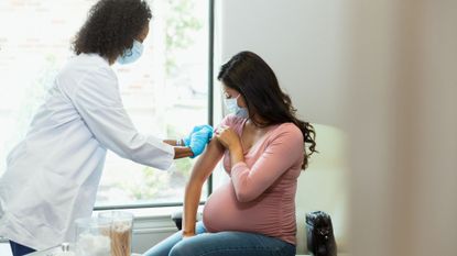 pregnant woman in a pink shirt sits in a doctor's office as her doctor puts a bandage on her arm after a vaccination