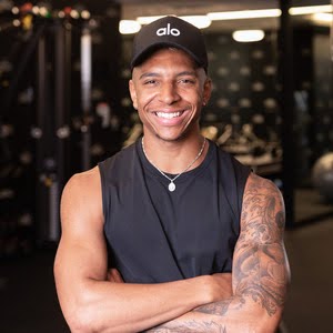 Smiling man with his arms crossed, wearing black tank top, pendent necklace and black cap with "alo" written on it in white text